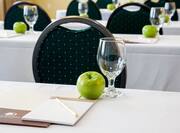 Detailed View of Meeting Room Tables With Green Apples, Drinking Glasses, Notepads, Pens on White Linens, and Chairs