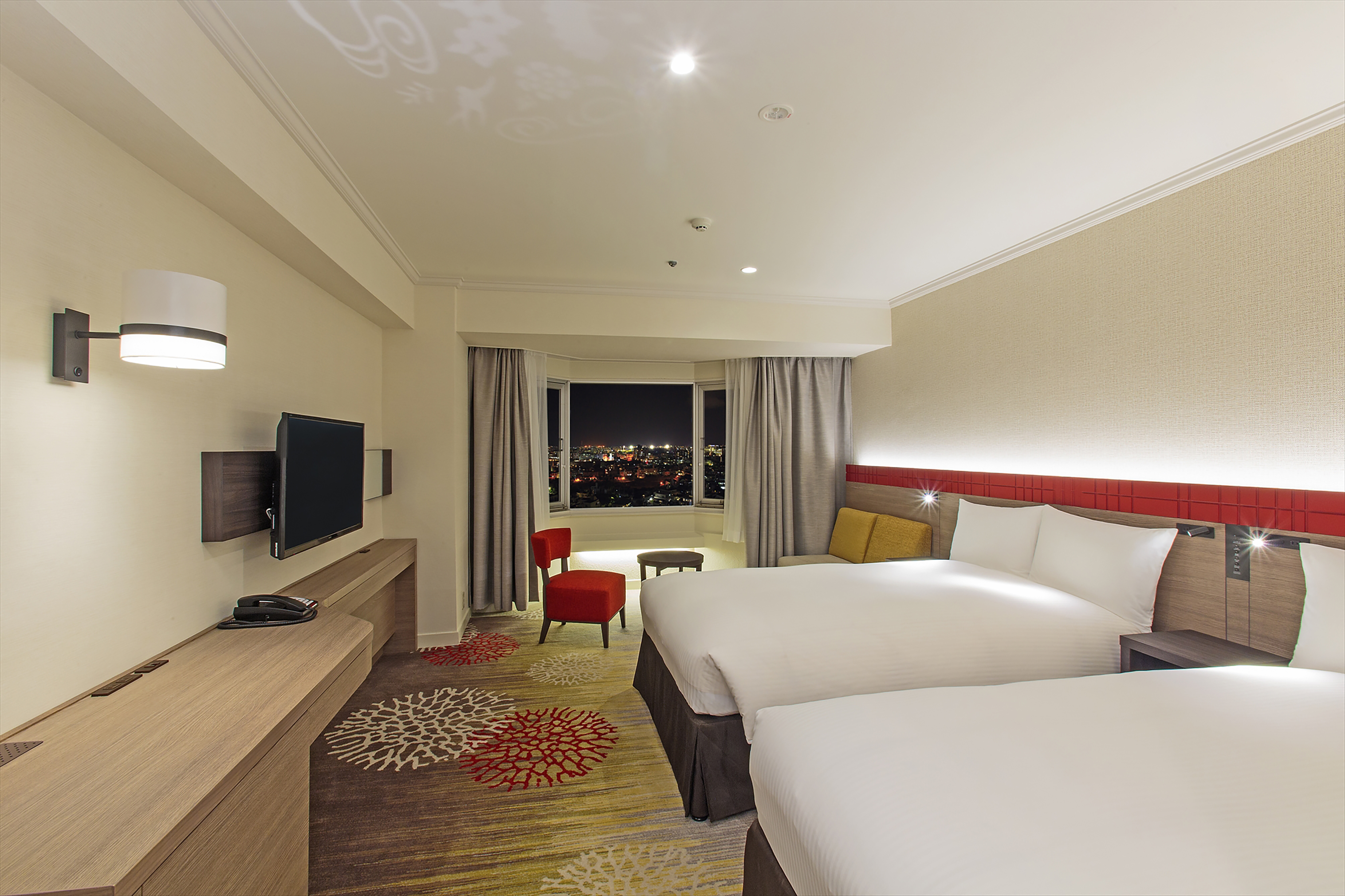Twin Beds, TV, Red Chair and Coffee Table By Window With Open Drapes to Illuminated City at Night in Premium Room