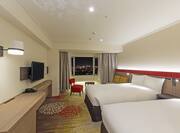 Twin Beds, TV, Red Chair and Coffee Table By Window With Open Drapes to Illuminated City at Night in Premium Room