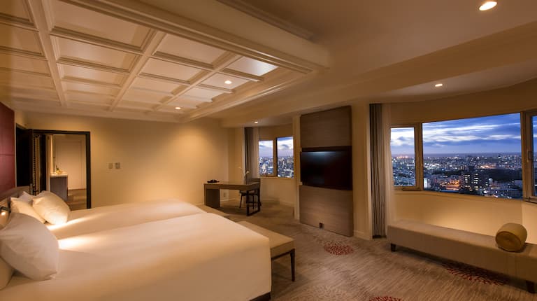 スイート客室内(ツインベッド2台、照明、リビングエリアに続くオープンドアウェイ、ワークデスク、照明、テレビ、ワークデスク、ネオンが灯った夕暮れ時の市街地を一望するオープンカーテン付きの窓、テレビ、ラウンジチェア)