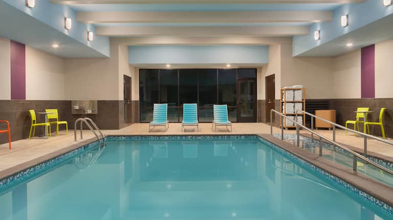 Illuminated Indoor Pool With Chairs, Loungers, Towel Station, Windows With Nighttime View 
