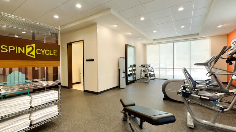Fitness Center With Signage Above Towel Station, Open Doorway to Laundry Center, Water Cooler, Large Mirror, Free Weights by Windows, Cardio Equipment and Weight Bench in Fitness Area of Spin2 Cycle