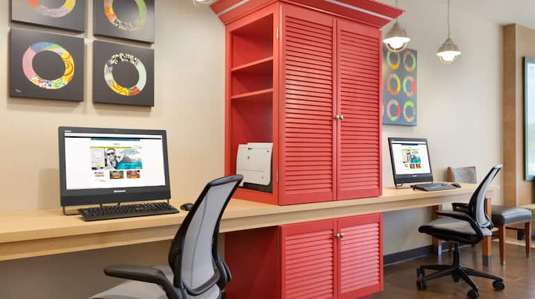 Wall Art, Red Storage Cabinet With Printer Between Two Computers on Long Desk, and Two Ergonomic Chairs in Business Service Area