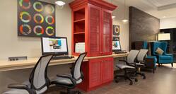 Business Center with Colorful Wall Art, Two Computers on Work Desk, Four Ergonomic Chairs, Red Storage Cabinet With Printer and Two Blue Armchairs by Illuminated Floor Lamp