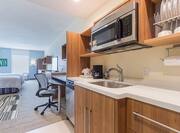 Kitchen With Dishwasher, Sink, Microwave, Dishes in Wood Cabinets, Queen Bed, Window, and Work Desk in Studio Suite