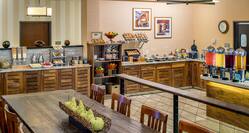 Long Wooden Table With Chairs, Wall Art, Wall Clock, Food Service Area With Plates, Utensils, Hot and Cold Breakfast Items