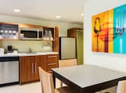 Wall Art Above Dining Table With Seating for Three, Kitchen With Dishes in Wood Cabinet, Dishwasher, Coffee Maker, Microwave, and Fridge in King Suite   
