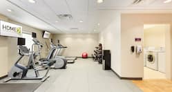  Spin2Cycle Fitness Area With Cardio Equipment Facing TV, Window,  Red Exercise Ball, Free Weights, Weight Balls, Water Cooler, and Open Doorway to Laundry Room With Coin Operated Washing and Drying Machines