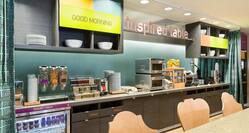Brightly Lit Signage Above Counter With Yogurt, Milk, Cereal, Condiments, Microwave, Fruit, Waffle Iron and Community Table in Inspired Table Dining Area