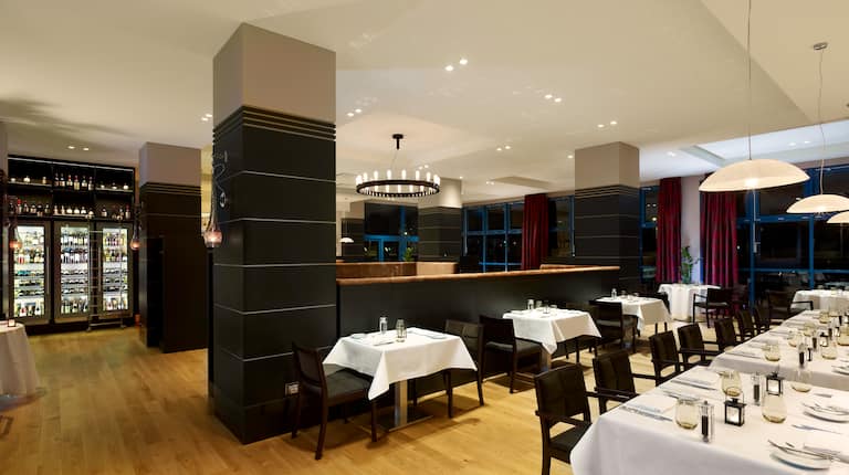 Prostřené stoly k večeři s výzdobou, bílými ubrusy, židlemi, stojanem na víno a okny s rozhrnutými závěsy s nočním výhledem v restauraci Laurus Mediterranean