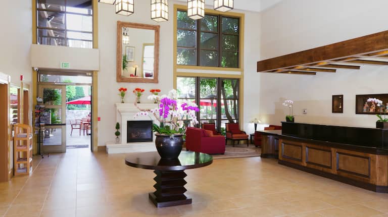 Decorative Lighting, Soft Seating by Fireplace, Glass Door and Windows With Patio View, Table With Flowers, and Front Desk in Lobby Lounge Area