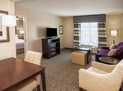 Living Room Area of Two Room Suite With Purple Sofa, Arm Chair, TV, Ottoman, Dining Table, and Open Doorway to Bedroom 