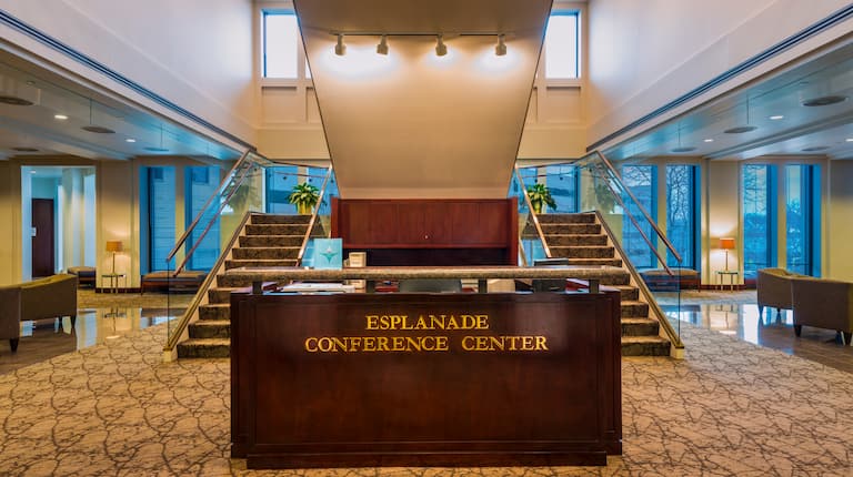 Esplanade Conference Center Signage on Reception Desk With View of Double Staircases, Soft Seating, and Windows in Background