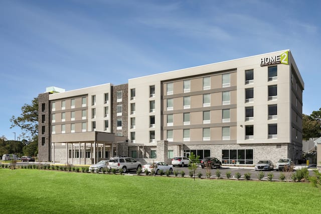 Modern Home2 Suites hotel featuring lush green lawn, ample parking, and bright blue sky.