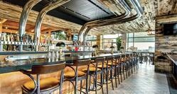 Image of a long empty bar with a row of wooden bar stools. industrial chrome pipes go from the back of the bar to the ceiling and rustic wood cladding is used in the decor.