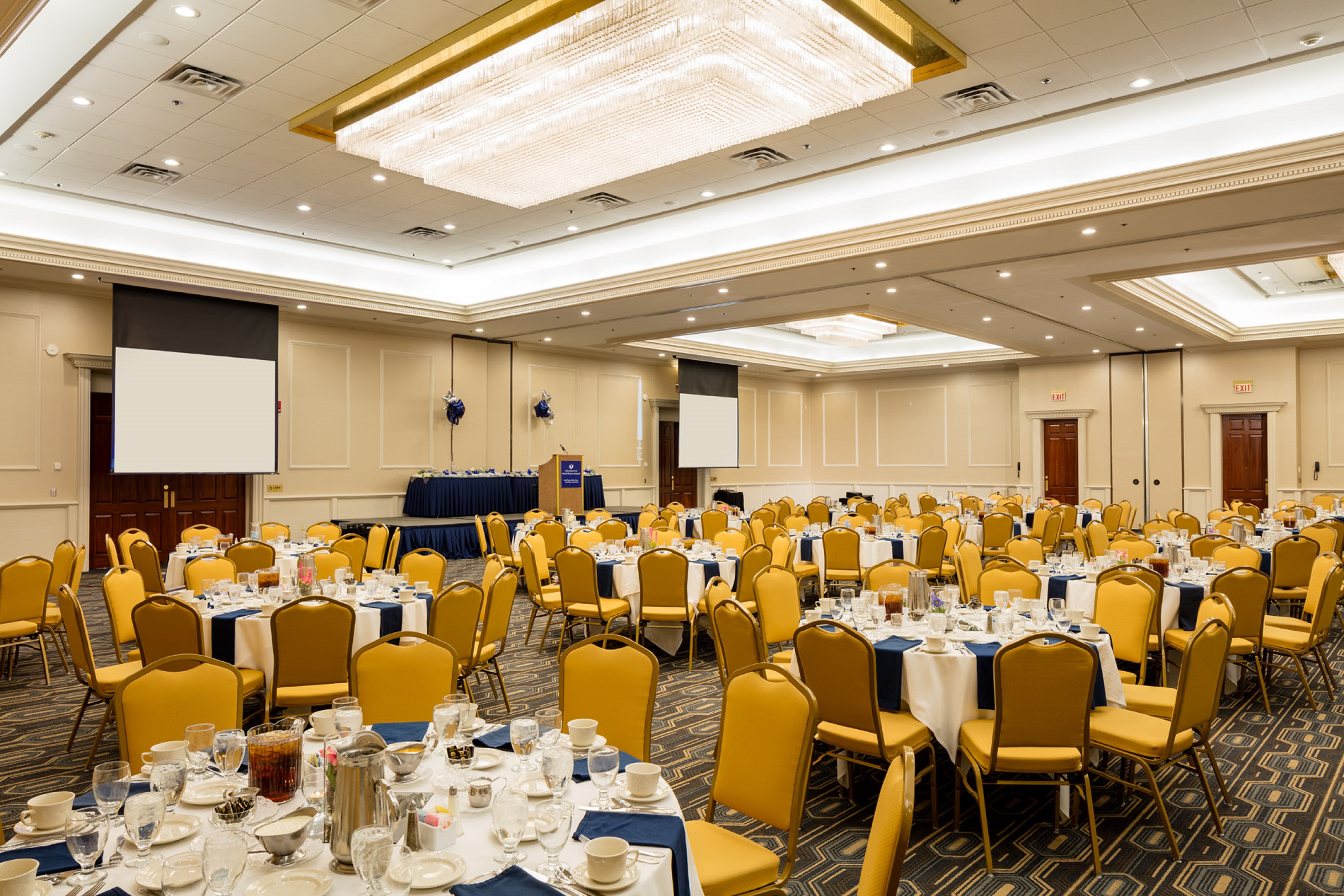 Ballroom Set Up for Special Event With Place Settings, Beverage Pitchers and Blue Napkins on Banquet Tables With White Linens, Yellow Chairs, Two Presentation Screen, and Stage With Podium