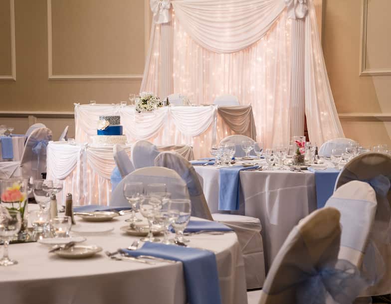 White Drapes Behind Head Table, Cake Table, and Detailed View of Dining Table With Place Settings, Flowers, Blue Napkins, White Table Number, and White Chairs With Blue Bows Set Up For Wedding Reception