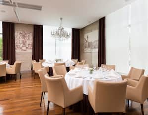 Paris Restaurant – Essbereich mit Gedecken, Blumen, Trinkgläsern und weißer Tischwäsche auf Tischen, beigen Stühlen, Fenstern mit offenen Gardinen und Parkblick, Wandbild und Stimmungsbeleuchtung