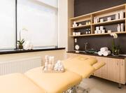 Spa Treatment Room With Towels and Toiletries on Massage Table, Window With Closed Shades, Sink and Fresh Flowers on Wood Cabinet