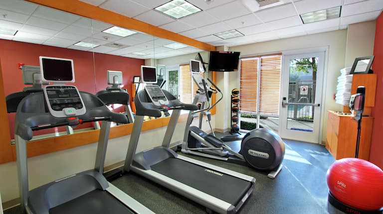 Fitness Center With Cardio Equipment Facing Mirrored Wall, TV, Weight Balls, Glass Entry Door, Towel Station, and Red Stability Ball