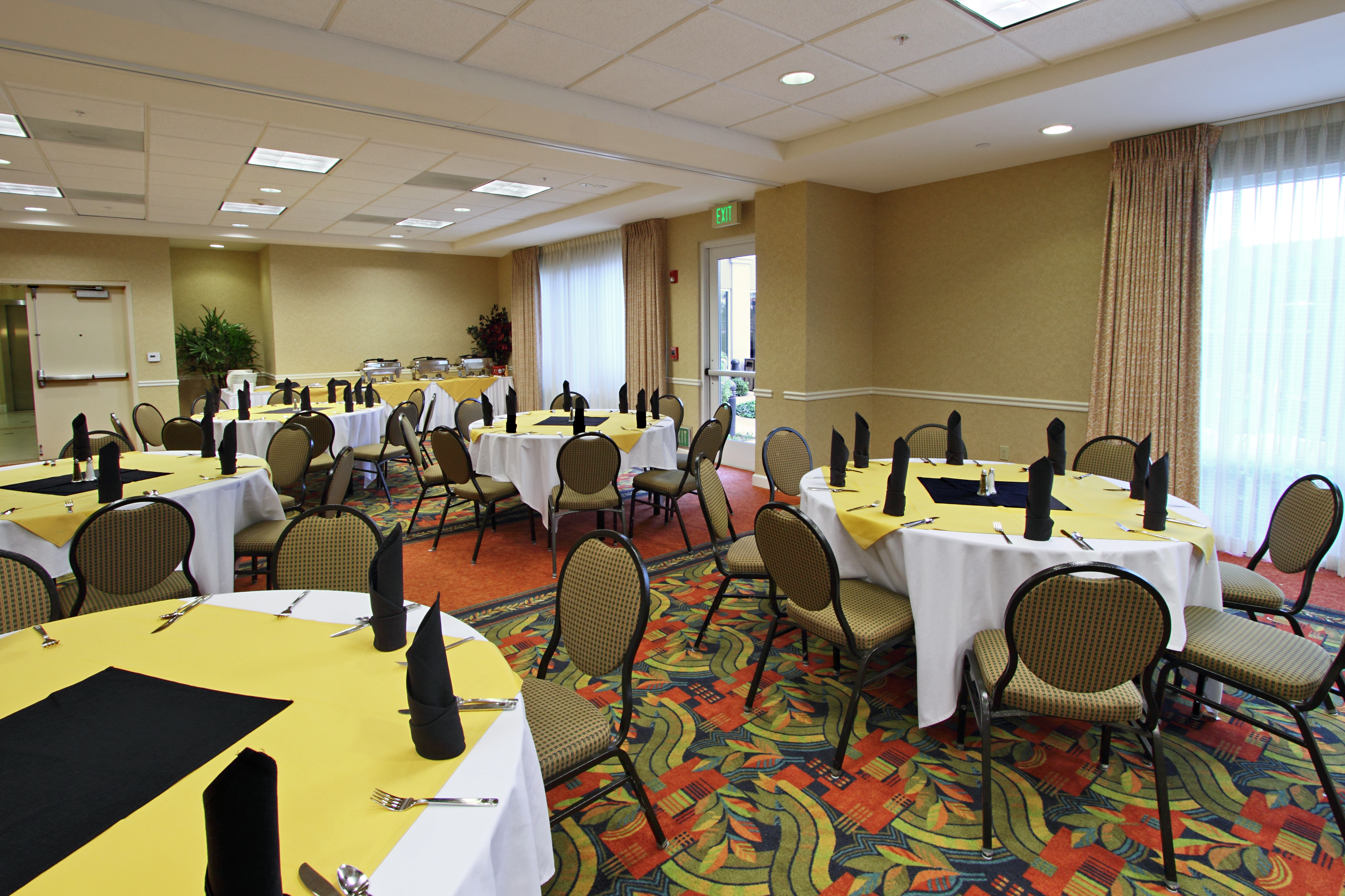 Silverware, Black Napkins, and Linens on Round Tables, Chairs, Food Service Area, Entry Doors and Windows With Long Drapes in Meeting Space Set up for Banquet
