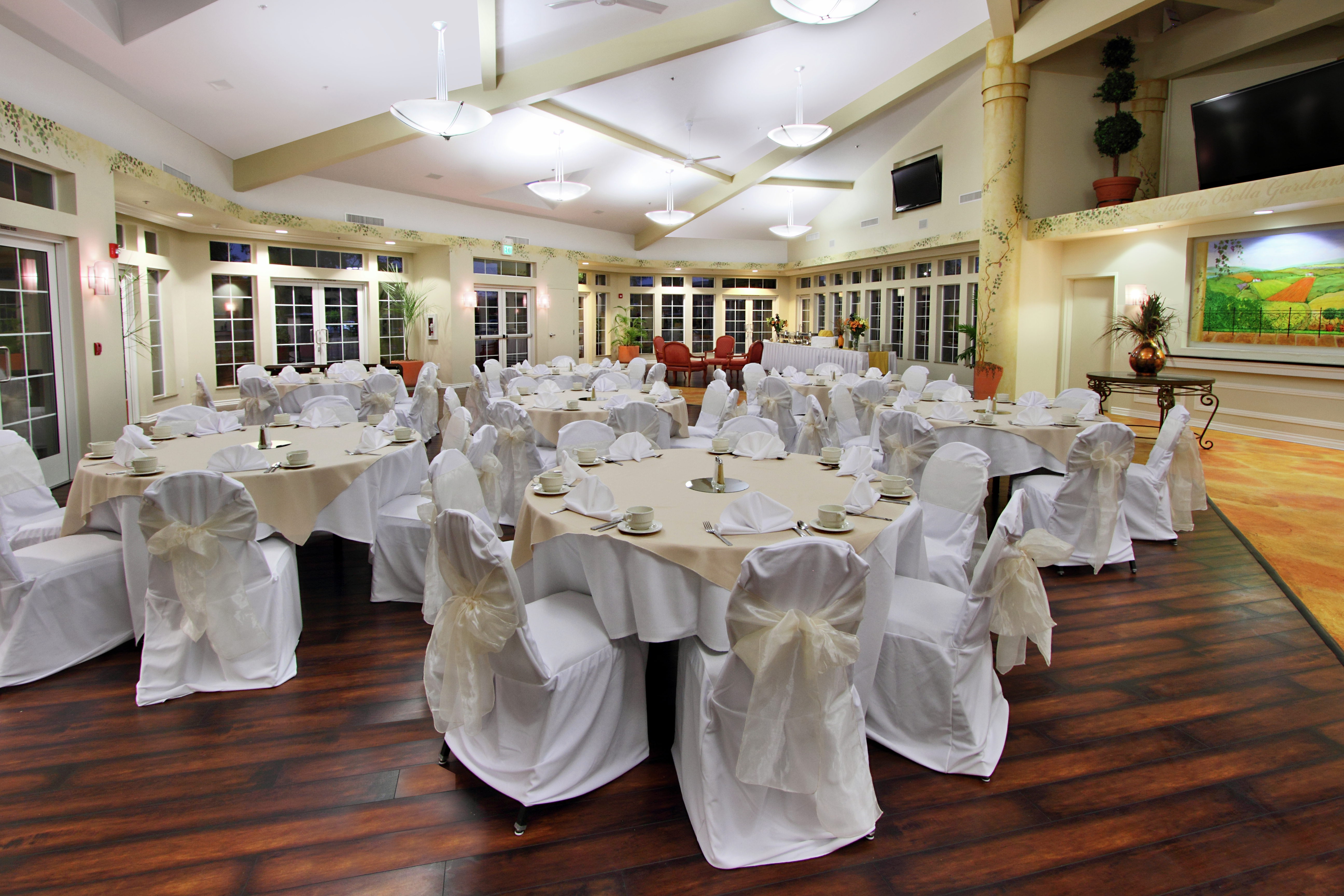 Place Settings and White Linens on Round Tables, White Chairs, TVs, and Large Windows in Meeting Space Set up for Wedding