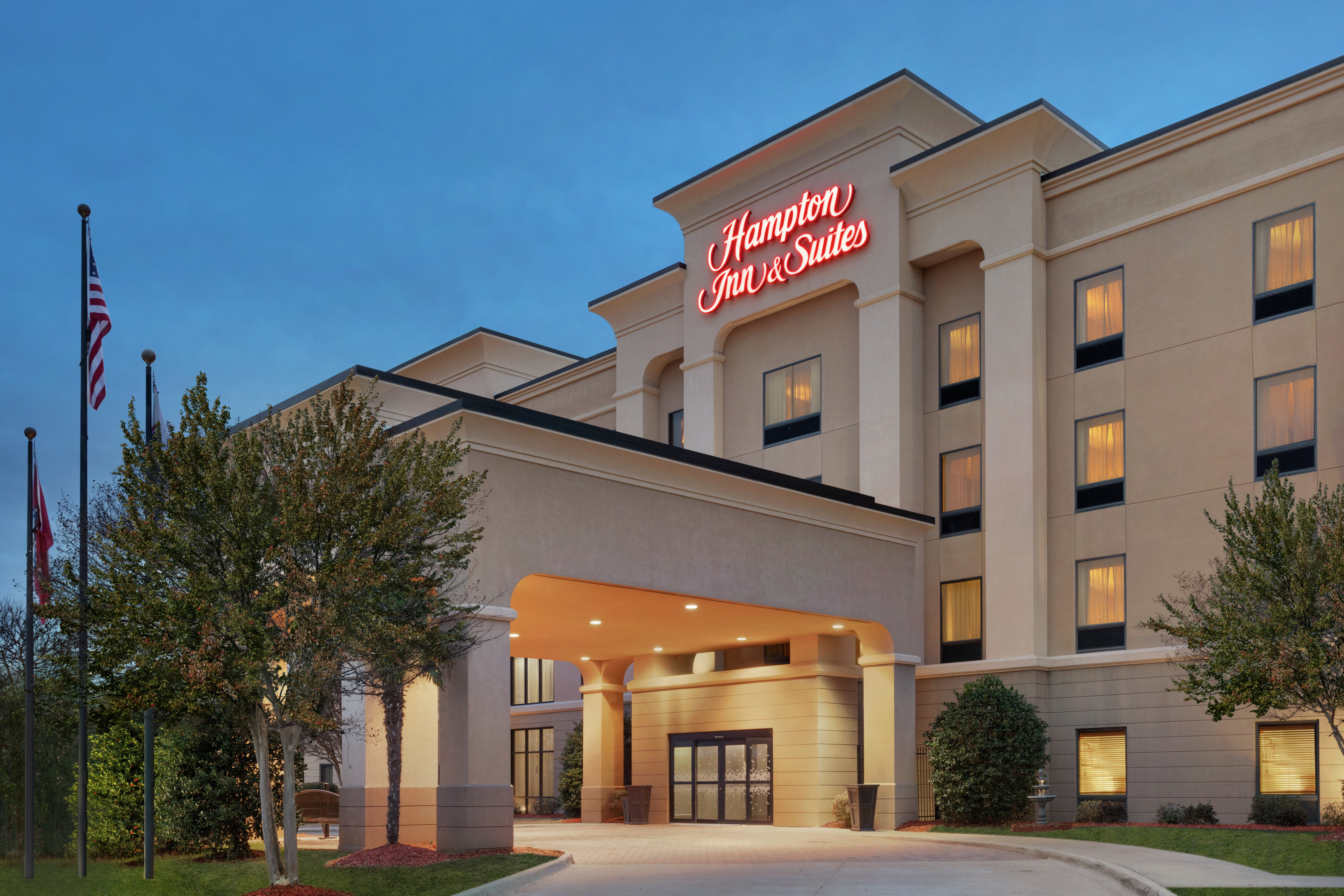 Welcoming Hampton Inn hotel exterior featuring covered entrance, lush landscaping, and glowing guestroom windows.