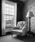 Кресло в стил „Честърфийлд“ с дизайн на цветята, което седи пред прозорец на саш, стояща лампа и тапет с флорално наследство