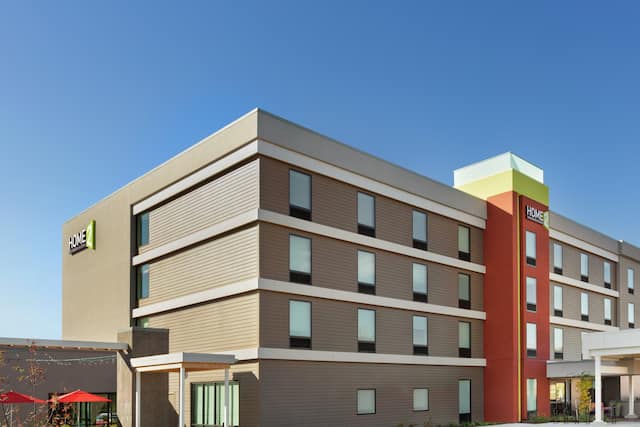 Modern Home2 Suites hotel exterior featuring porte cochere, patio with umbrellas, and bright blue sky.