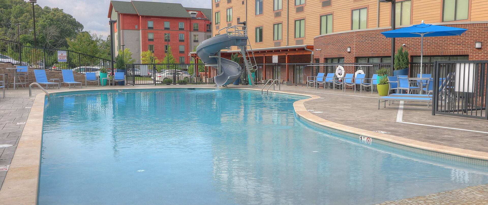 Daytime View of Leisure Seating, Water Slide, and Table With Sun Umbrella by Outdoor Pool and Hotel Exterior