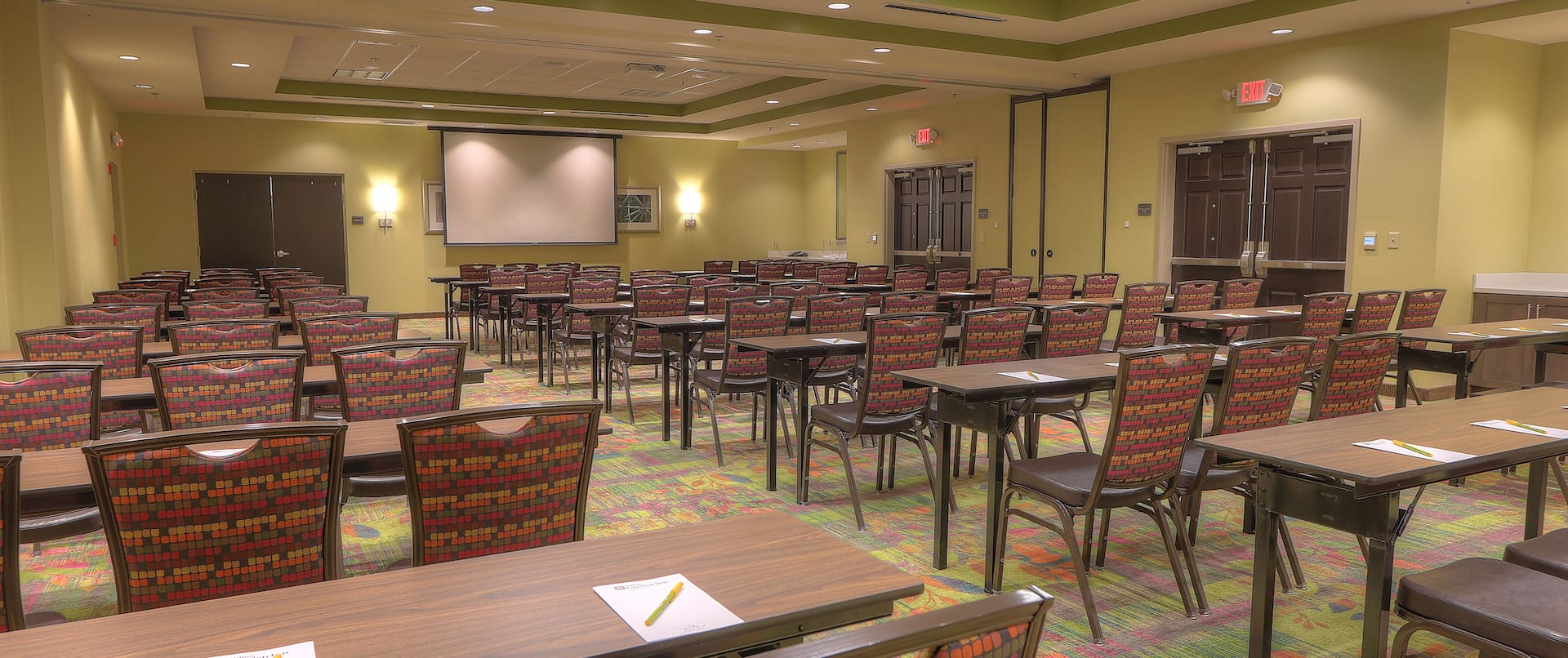Classroom Setup in Meeting Room With Notepads and Pens on Tables, Chairs, Presentation Screen, Overhead Projector, Wall Art, and Entry Doors
