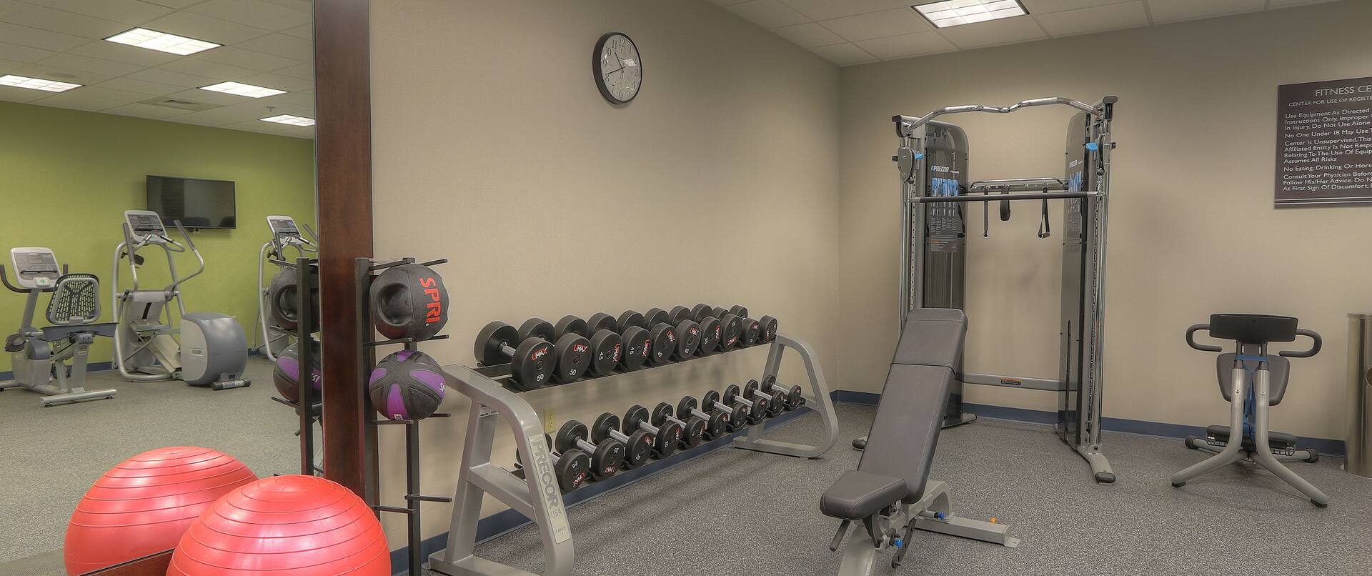 Cardio Equipment and TV Reflected in Large Mirror, Red Exercise Ball, Weight Balls, Free Weights, Wall Clock, Weight Machine, and Weight Bench in Fitness Center