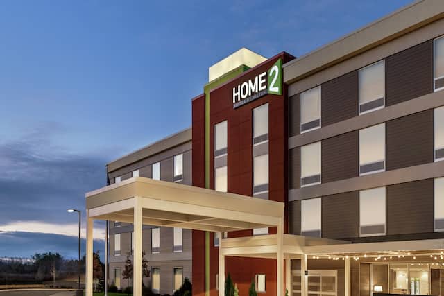 Modern Home2 Suites hotel exterior featuring porte cochere, glowing outdoor patio, and beautiful dusk sky.