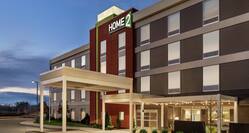 Modern Home2 Suites hotel exterior featuring porte cochere, glowing outdoor patio, and beautiful dusk sky.