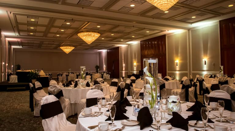 Flowers in Water With Floating Candles, Place Settings, and Black Napkins on Tables With White Linens, and White Chairs With Black Bows in Ballroom Set Up for Wedding