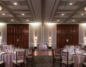 Place Settings, White Napkins, and Drinking Glasses on Dining Tables With Pink Linens and Dance Floor Set Up in Meeting Space For Banquet