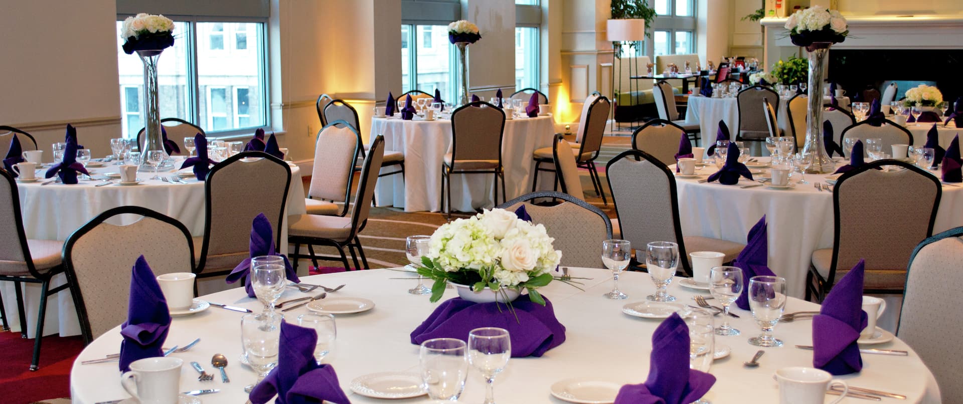 Fireplace in Meeting Room With Flowers, Place Settings, and White Linens on Round Tables Set Up For Daytime Banquet