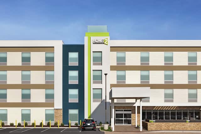 Modern Home2 Suites hotel exterior featuring large parking lot, outdoor patio, and beautiful blue sky.