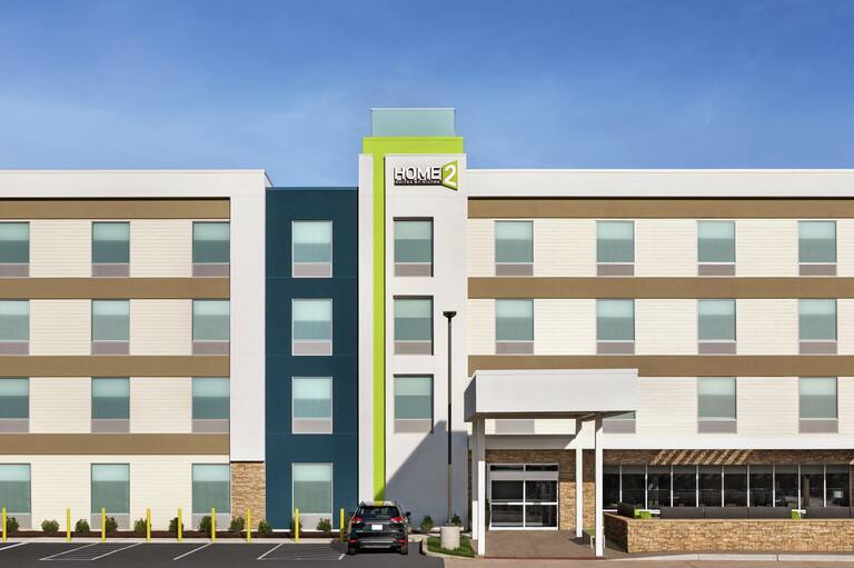 Modern Home2 Suites hotel exterior featuring large parking lot, outdoor patio, and beautiful blue sky.