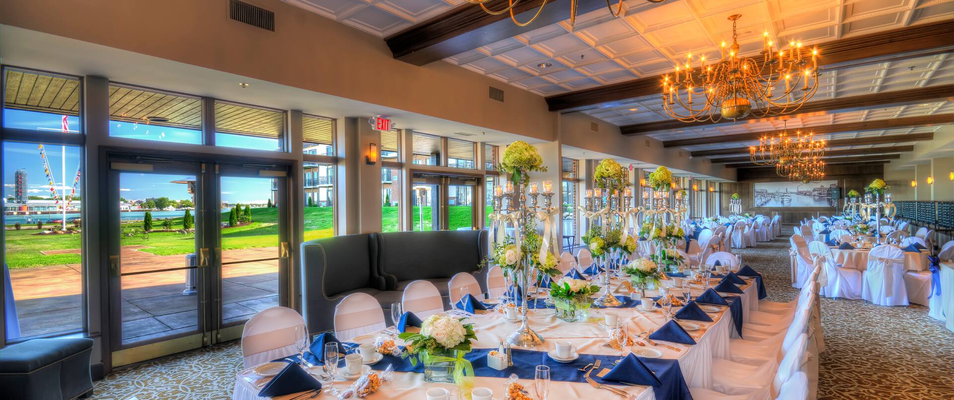 Glass Doors and Windows With Views, Head Table With Place Settings, Flowers, Blue Napkins, and Candles on White Linens, White Chairs, Soft Seating in Banquet Room