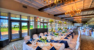 Glass Doors and Windows With Views, Head Table With Place Settings, Flowers, Blue Napkins, and Candles on White Linens, White Chairs, Soft Seating in Banquet Room