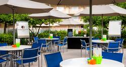 Tables With Sun Umbrellas and Blue Chairs, Two Easels, and Beverage Table Set Up for Event in Courtyard