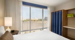 Neatly Made Bed, Illuminated Lamp on Bedside Table by Large Window With Stadium View, Closet With Blue Blue Drapes and Wood Shelves