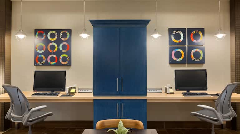 Business Center With Blue Storage Cabinet Between Two Computers on Long Desk, Two Ergonomic Chairs, and Colorful Wall Art, 