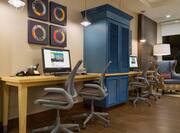  Business Service Area With Colorful Wall Art, Blue Storage Cabinet Between Two Computers on Long Desk, and Four Ergonomic Chairs, Arm Chair and Illuminated Floor Lamp by Window