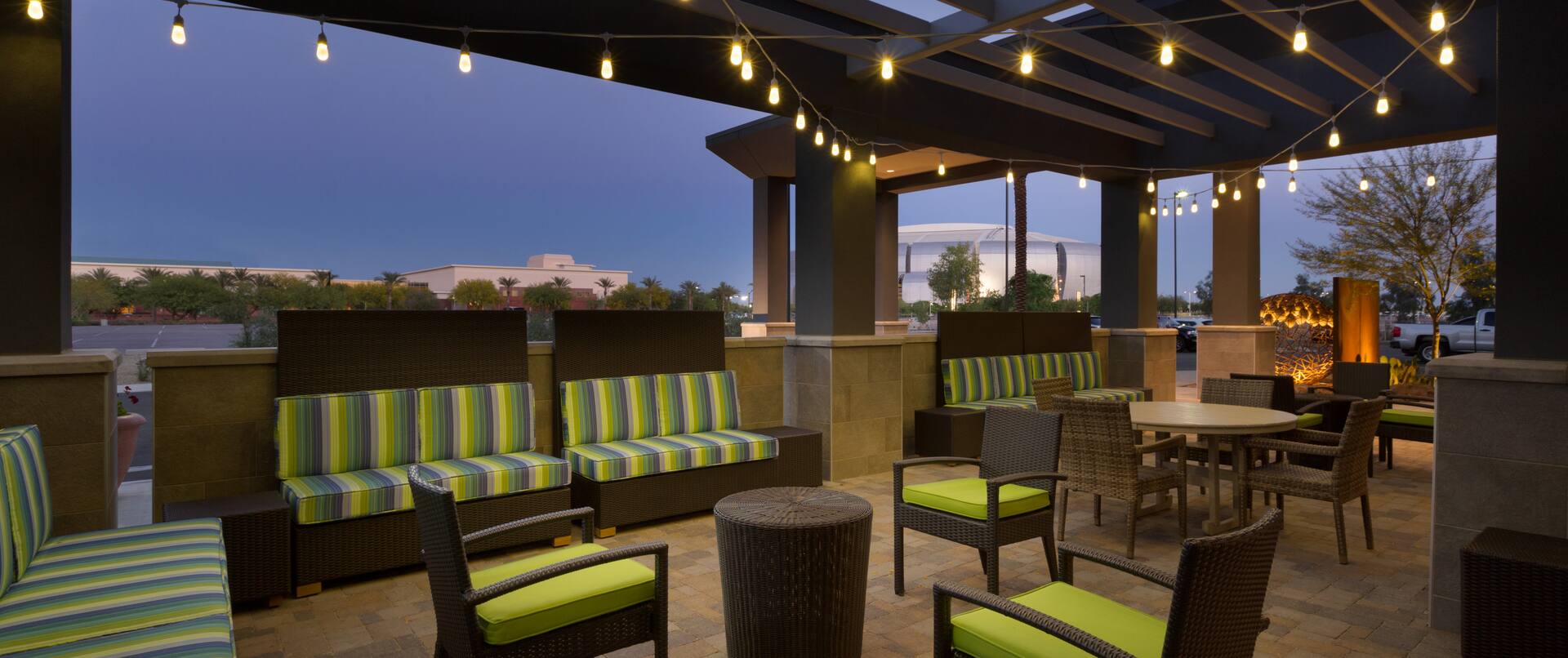 Round Table, Armchairs With Green Cushions and Striped Sofas Under Pavilion With Illuminated String Lights on Outdoor Patio at Dusk