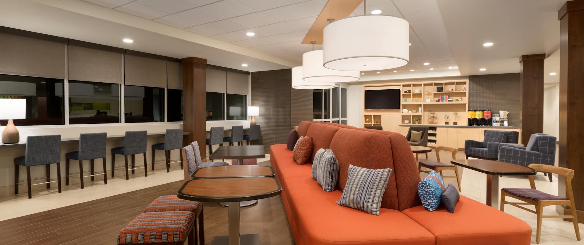 Oasis Lounge in Lobby With Table Seating by Large Orange Sofa, Decorative Lighting, Counter Seating by Windows, and View of TV and Beverage Station in Background