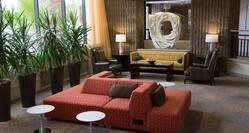 Art Feature Surrounded by Soft Seating, Tables, Illuminated Lamps, Plants and Large Windows With Open Drapes in Lobby