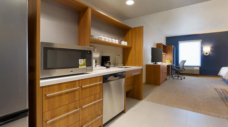 Accessible Studio Suite With Fridge, Microwave on Counter, Dishes in Wood Cabinets, Coffee Maker on Counter, and Dishwasher in Kitchen, TV, Work Desk, Illuminated Lamp by Window and Queen Bed