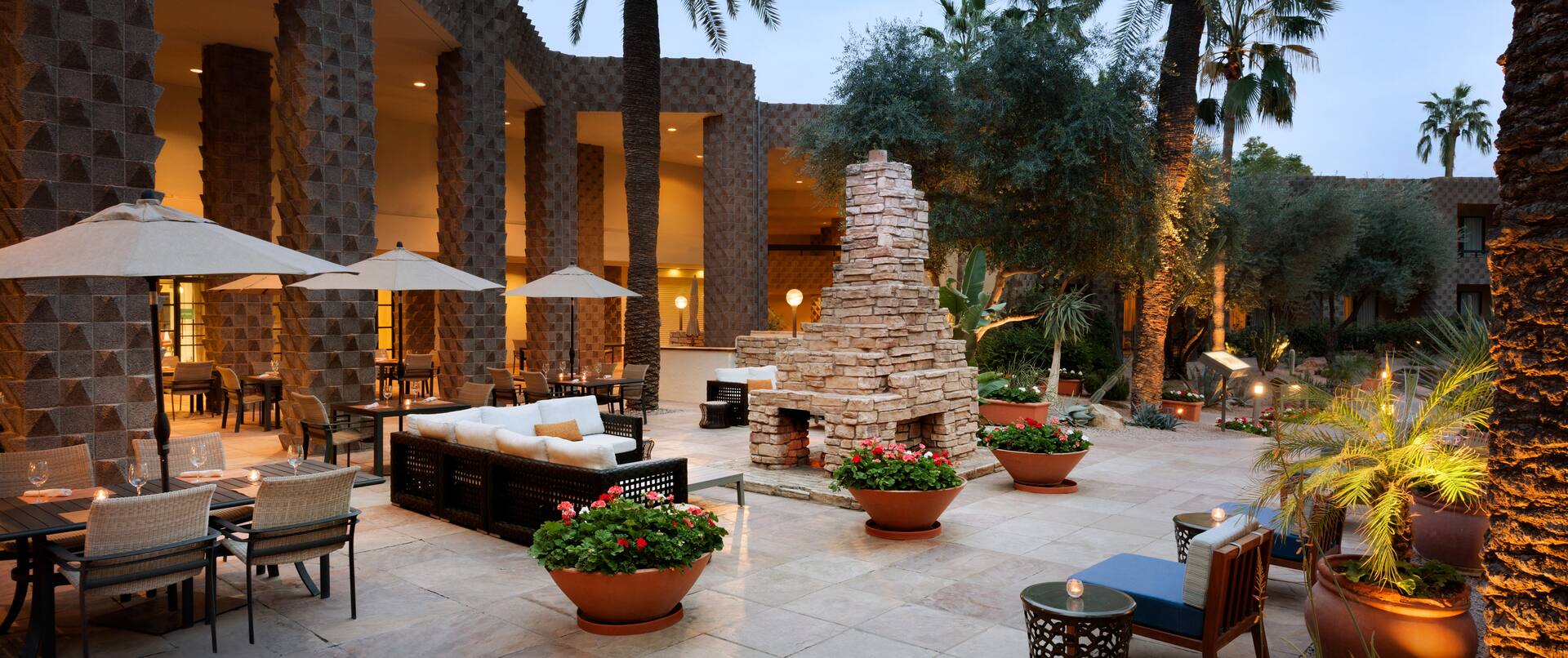 Tables With Umbrellas, Soft Seating, Furnace, and Candlelit Tables on Outdoor Patio Surrounded by Palm Trees at Dusk